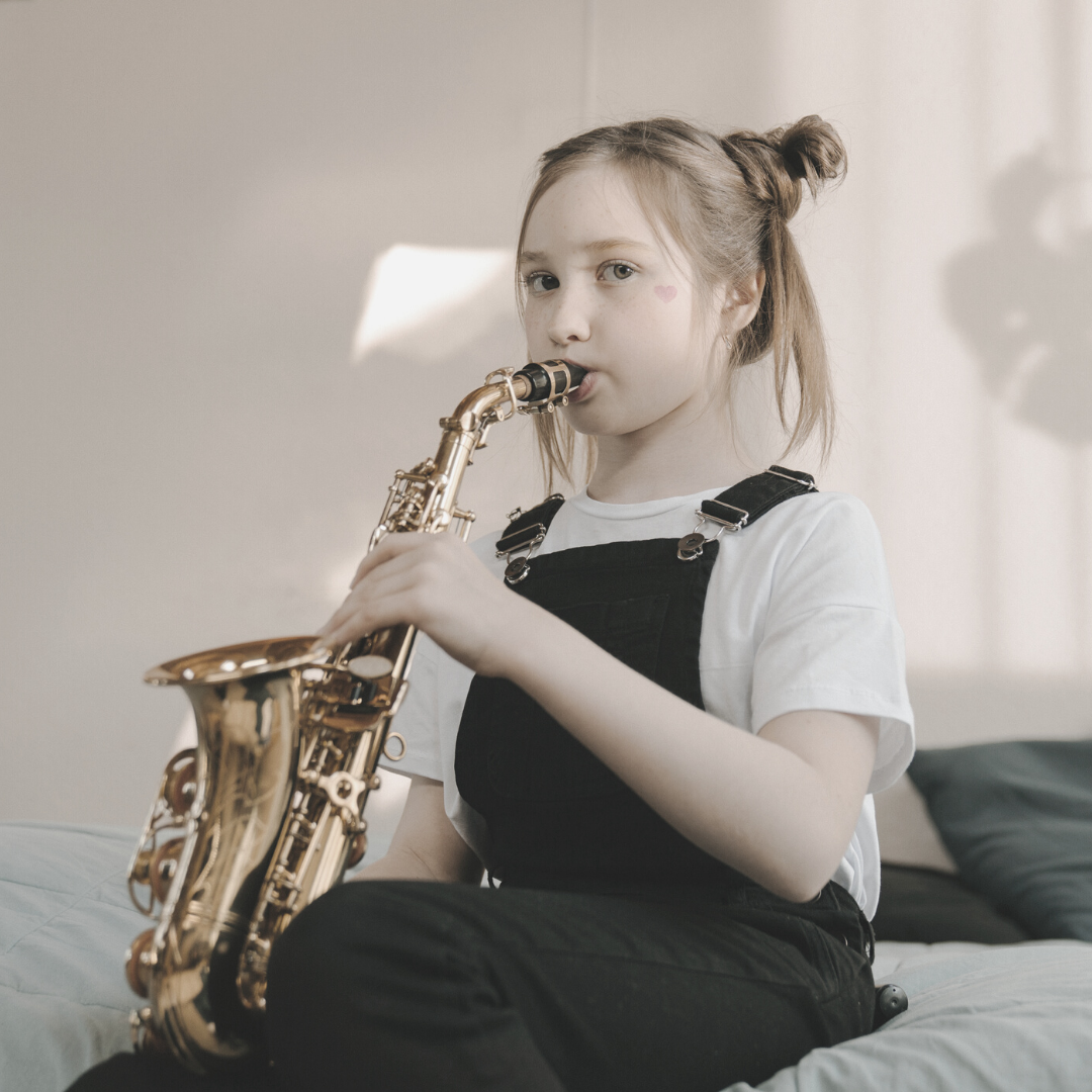 young girl wearing dungarees sat on bed playing saxophone